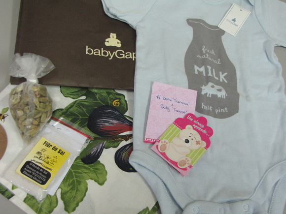 foto de um macacão de bebê, um saquinho com sementes de cardamomo, um saquinho de flor de sal e um pano de mão com desenhos de figos